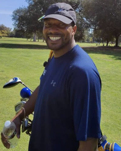 Chandler smiling on a golf course in a dark cap, carrying his golf bag
